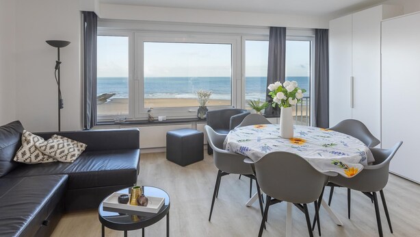 A modern living room with a sofa, dining table, and chairs, large windows with a beach view, and a ceiling light fixture. - Cosy studio with sleeping area and sea view (Middelkerke)