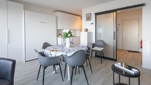 A modern, well-lit dining area with a round table, grey chairs, and a vase of flowers. There is a small side table with decorative items. - Cosy studio with sleeping area and sea view (Middelkerke)