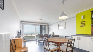 Una sala de estar moderna con una mesa redonda de madera y sillas, un sofá, un ventanal con vistas a la playa y una pared de acento de color amarillo brillante.