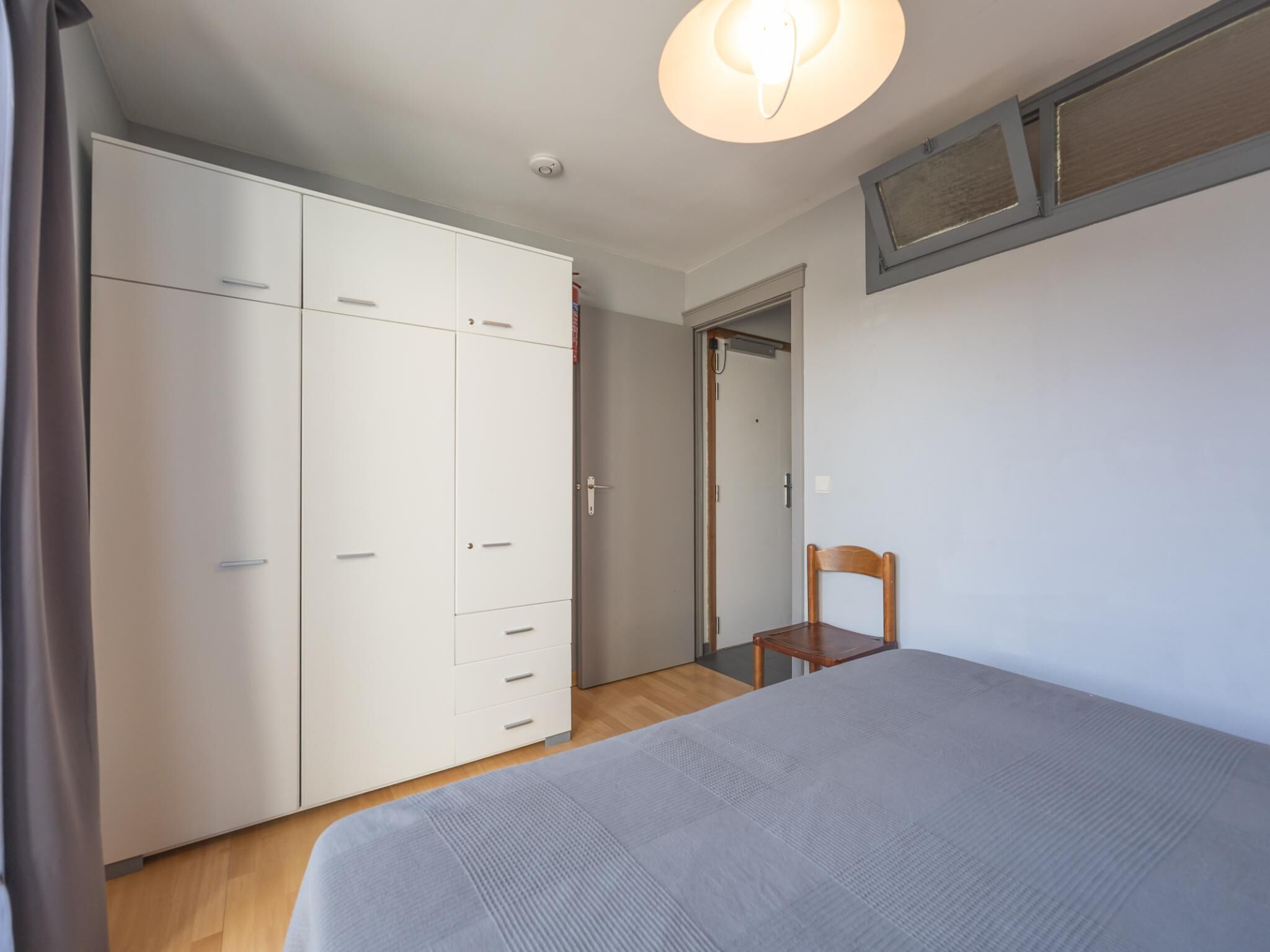 Et soveværelse med seng, garderobe, stol og en dør til et andet rum.