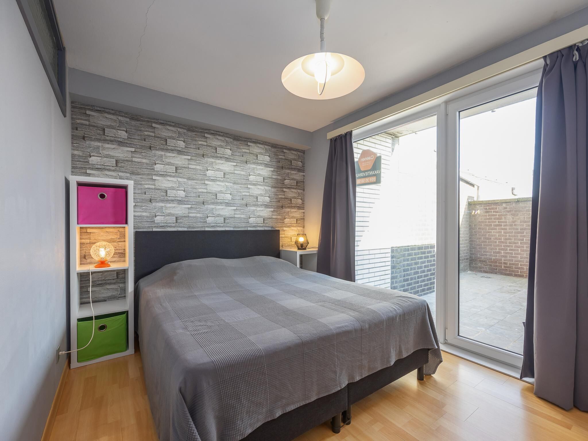 A modern bedroom with a large bed, a stone wall, a window with curtains, and a small table with a lamp.