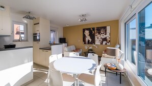 Eine moderne Wohnung mit Küche, Essbereich und Wohnraum. Sie verfügt über ein großes Fenster, einen Flachbildfernseher und einen Esstisch mit Stühlen.