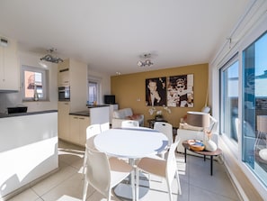 Un appartement moderne comprenant une cuisine, un coin repas et un espace de vie. Il est doté d’une grande fenêtre, d’un téléviseur à écran plat et d’une table à manger avec des chaises.