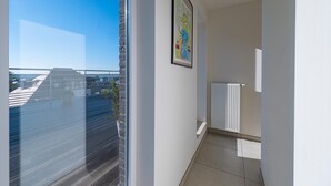 Ein schmaler Flur mit einem Fenster, das auf eine Stadtansicht blickt, einem Heizkörper und einem Bilderrahmen mit einem abstrakten Gemälde an der Wand.