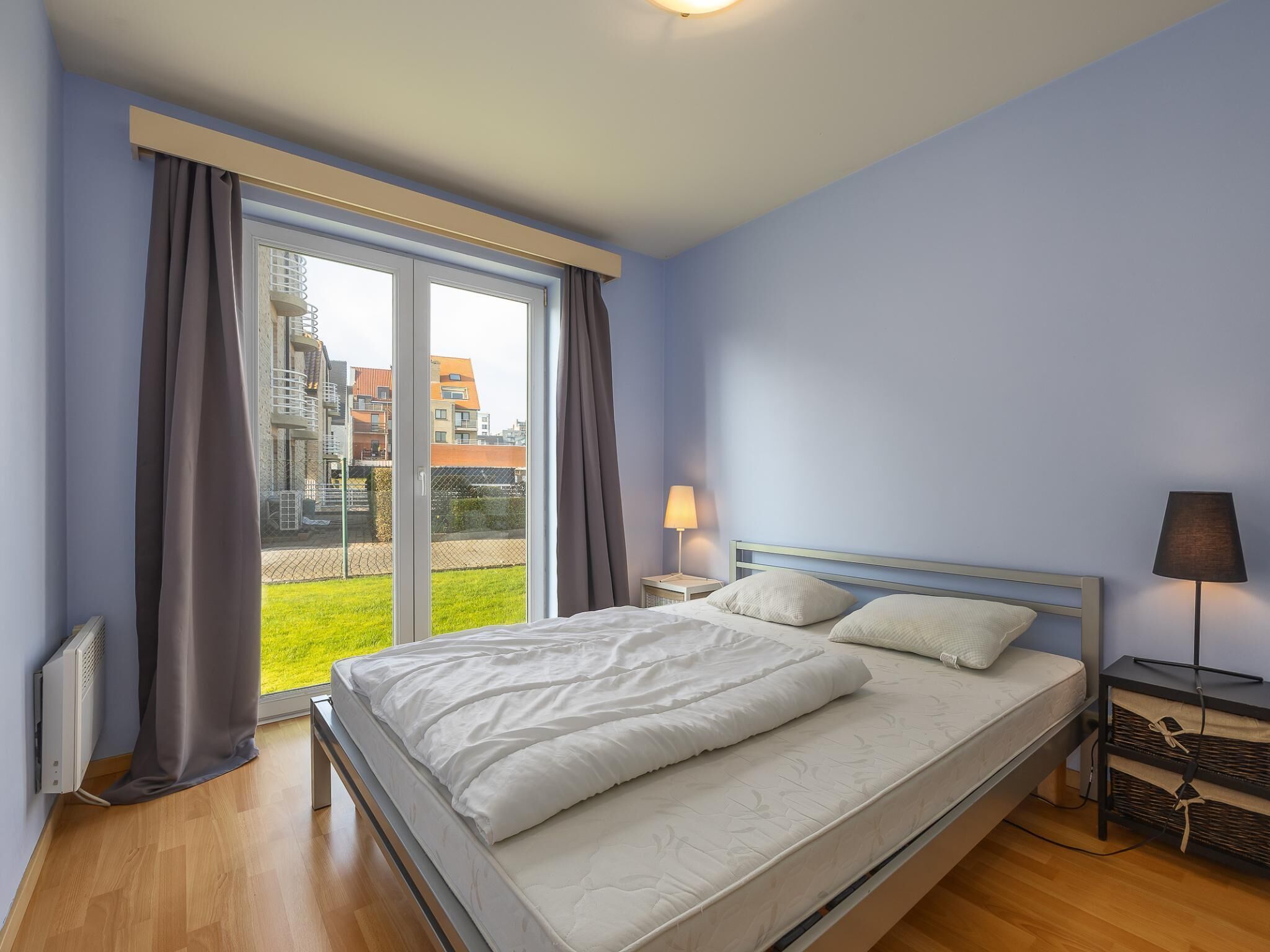A bedroom with a large bed, a nightstand, a lamp, and a view of buildings through a window.