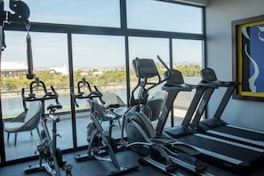 A gym with various exercise equipment, including stationary bikes and treadmills, with a view of a cityscape through large windows. - Sunrise & View (Mazatlán)