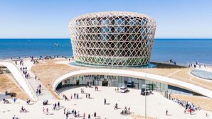 A modern architectural building with a unique geometric design, situated by a waterfront with a promenade and people walking around. - Apartment on the 6th floor with sea view (Middelkerke)