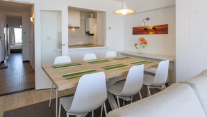 A modern dining area with a wooden table and white chairs, a kitchen in the background, and a painting on the wall. - Stunning apartment with phenomenal sea view (Middelkerke)