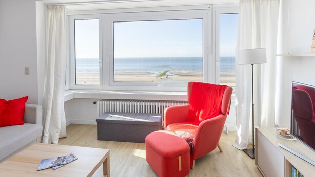 A room with a large window overlooking a beach, a red chair, a small table, and a radiator. - Stunning apartment with phenomenal sea view (Middelkerke)