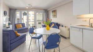 Un soggiorno moderno con un divano blu, un tavolo da pranzo rotondo con sedie, una zona cucina con pensili bianchi e vista attraverso le porte vetrate.