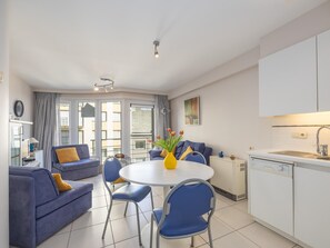 Ein modernes Wohnzimmer mit einem blauen Sofa, einem runden Esstisch mit Stühlen, einer Küchenzeile mit weißen Schränken und Blick durch Glastüren.