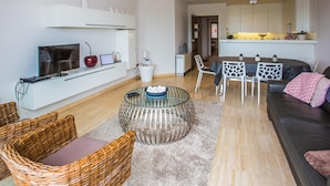 A modern living room with a TV, laptop, and decorative items on a white console. There is a glass coffee table, wicker chairs, and a sofa with a pink throw. A kitchen area is visible in the background. - Cozy apartment near the sea (Middelkerke)