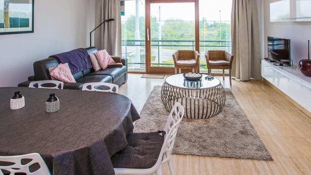 A modern living room with a dining table, sofa, and chairs. There is a large window with a view of greenery outside. - Cozy apartment near the sea (Middelkerke)