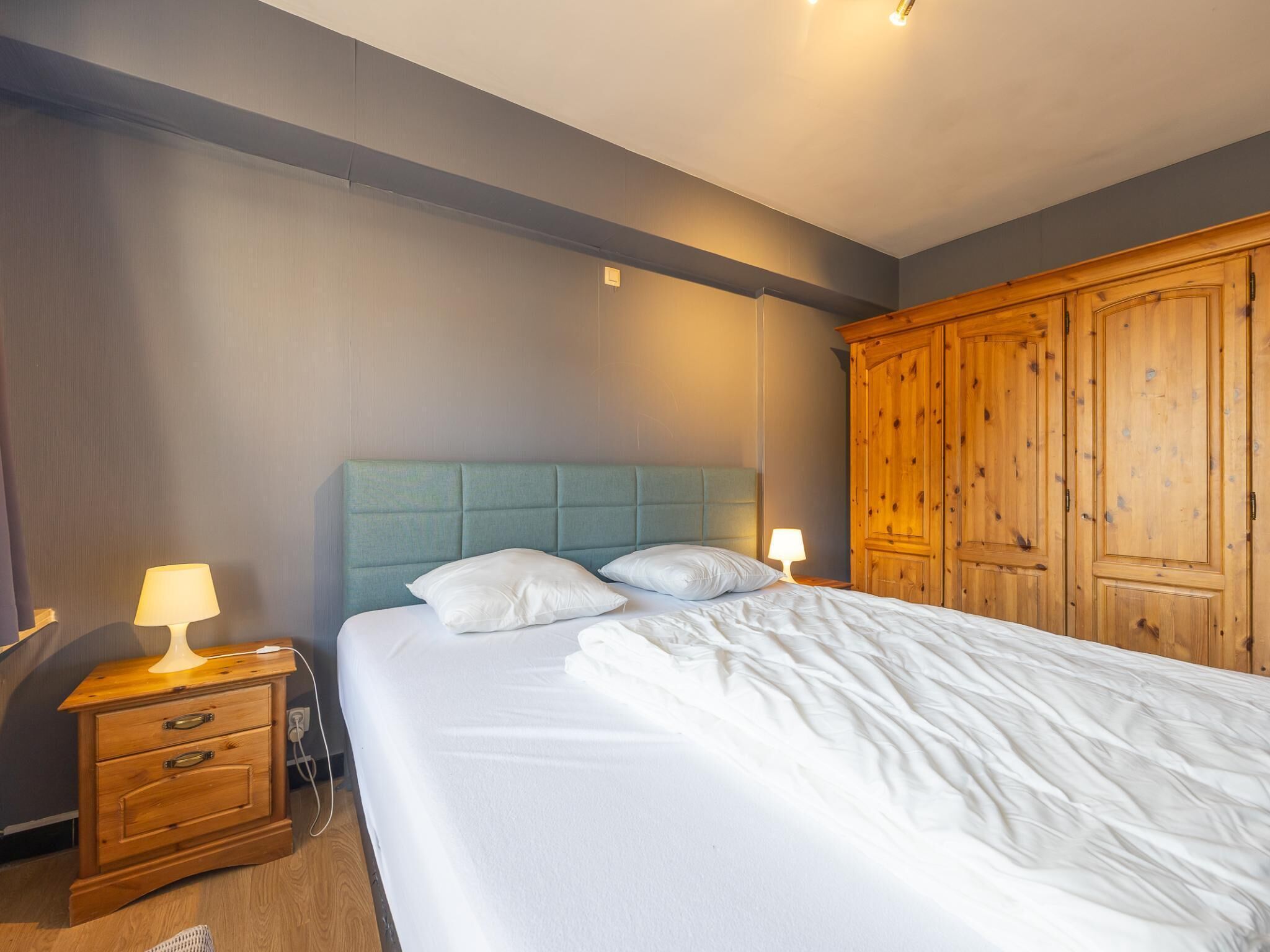Un lit bien fait, avec du linge de lit blanc et une tête de lit bleue, une table de chevet en bois avec une lampe, et une armoire en bois.
