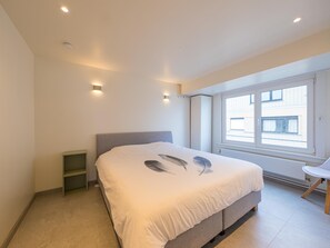 Un dormitorio con una cama grande, una mesita de noche pequeña, una silla y una ventana con vistas a edificios.