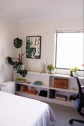 Ein Schlafzimmer mit einem Bett, einem weißen Schreibtisch, einem Bücherregal, einem Fenster und einer Topfpflanze.