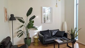 Een ruime woonkamer met een leren bank, een salontafel en een vloerlamp. Er staan verschillende potplanten en er hangt een schilderij aan de muur.