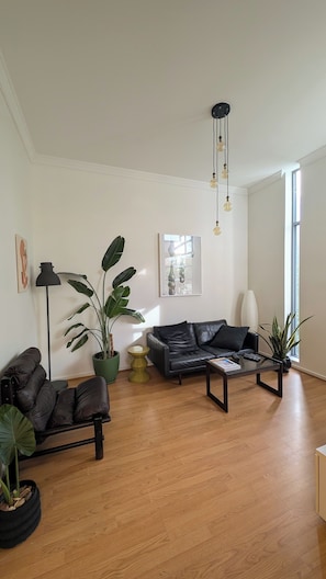 Ein geräumiges Wohnzimmer mit einer Ledersofa, einem Couchtisch und einer Stehlampe. Mehrere Blumentöpfe und ein Gemälde zieren die Wand.