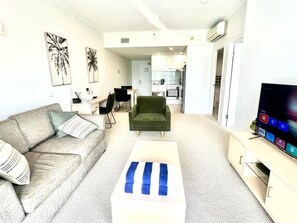Ein modernes Wohnzimmer mit einer Couch, einem Couchtisch, einem Fernseher und einer Küche im Hintergrund.