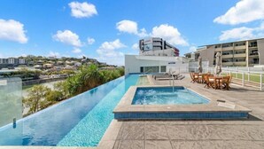 Une piscine à débordement sur le toit, offrant une vue sur le paysage urbain, un bâtiment portant l’inscription « Adani », et des espaces de détente en plein air.