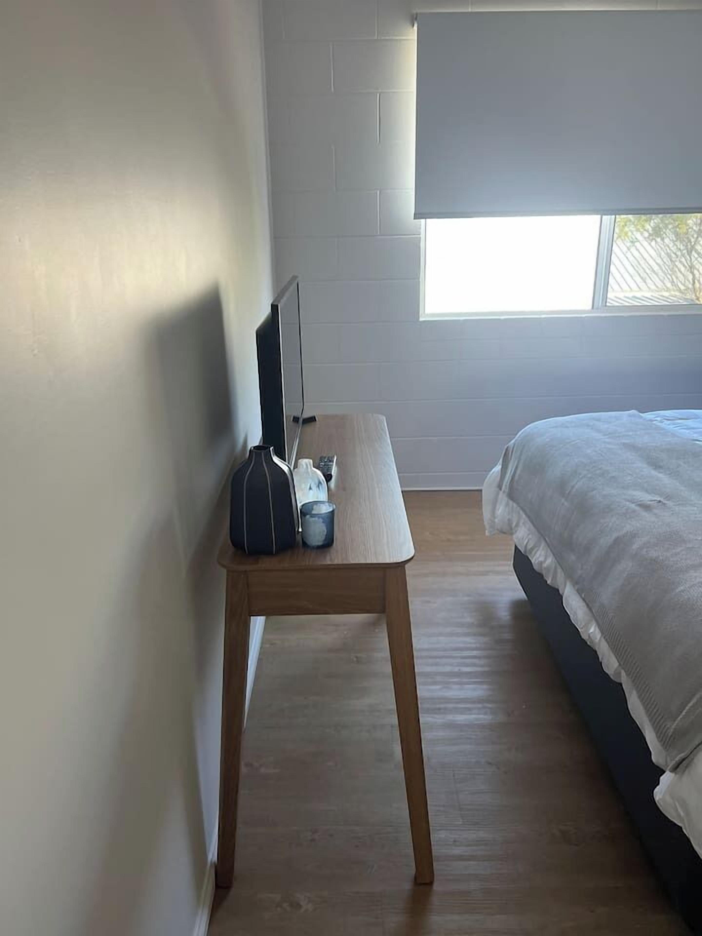 A bedroom with a bed, a bedside table with a lamp, a window, and a wall-mounted TV.