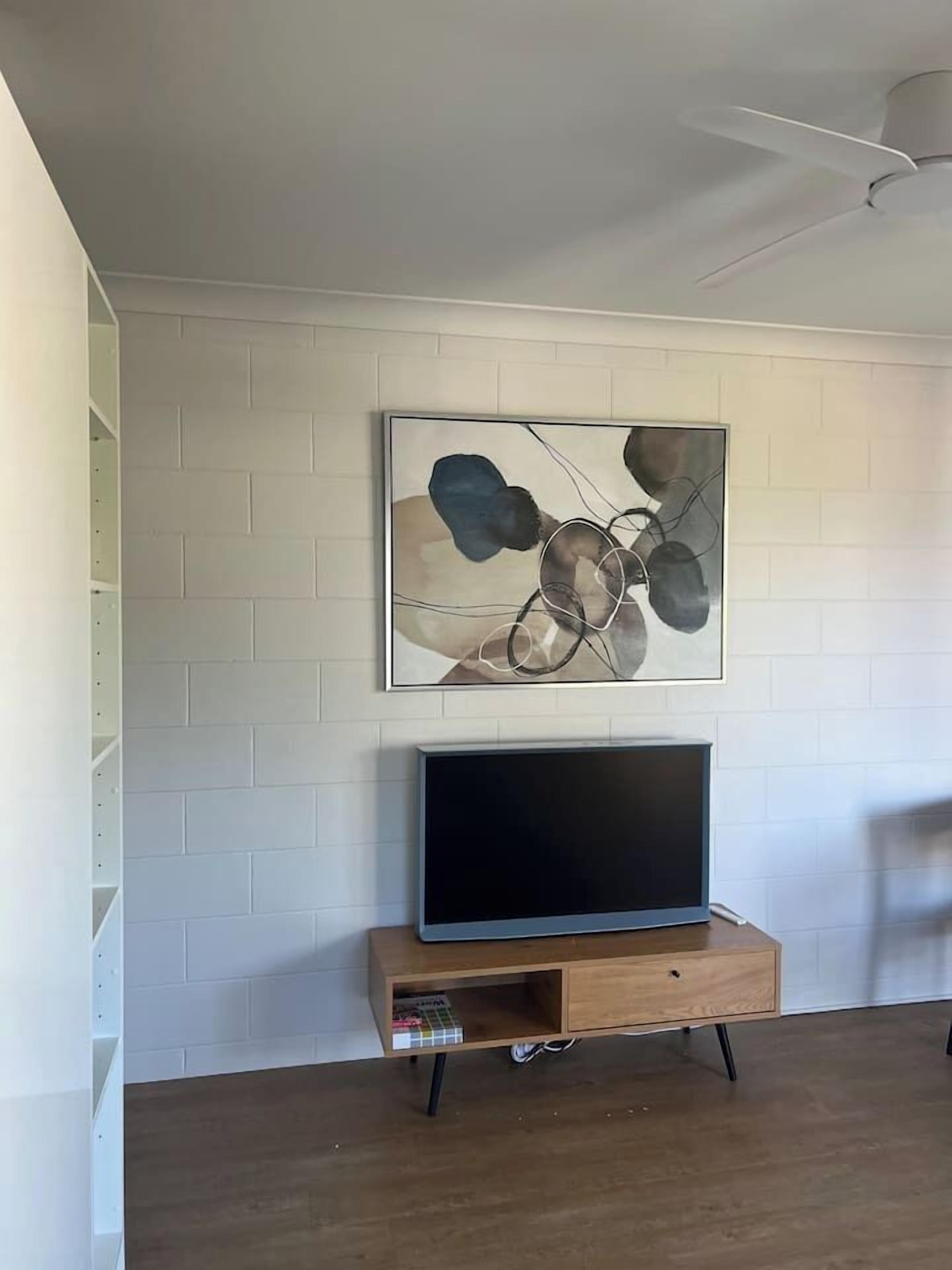 Une pièce avec un mur en briques blanches, un téléviseur à écran plat posé sur un meuble en bois et un ventilateur de plafond.