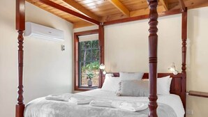 A four-poster bed with a white bedspread, a potted plant on the windowsill, and a wall-mounted air conditioner.