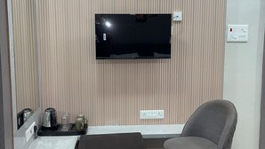 A modern hotel room with a flat-screen TV mounted on the wall, a small table with a kettle and glasses, a grey armchair, and a trash bin. - Hotel R J Heights (Fatehpur)