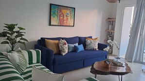 Een blauwe bank met bloemenkussens, een groen gestreepte stoel, een houten bijzettafeltje met een plant, en een schilderij aan de muur.