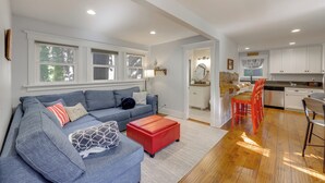 Un salon moderne avec un canapé gris, un pouf rouge et un coin cuisine doté de meubles blancs et d’un plancher en bois.