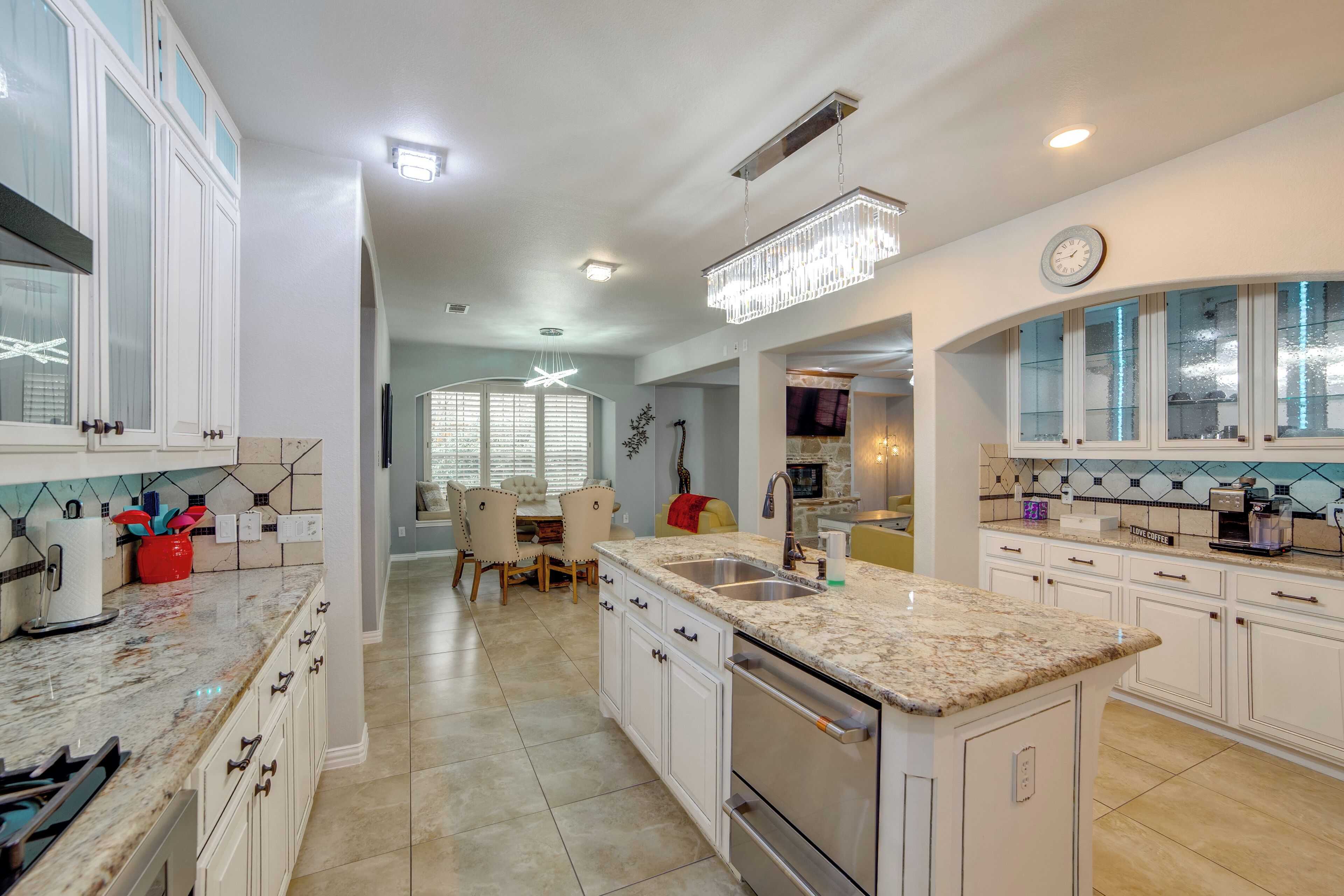 A modern kitchen with white cabinetry, granite countertops, and stainless steel appliances. There is a dining area with a table and chairs, and a chandelier hanging above.