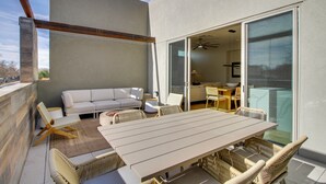 En balkon med et træbord og fletstole, en hvid sofa og en glasdør, der fører ind til et område med ventilator.