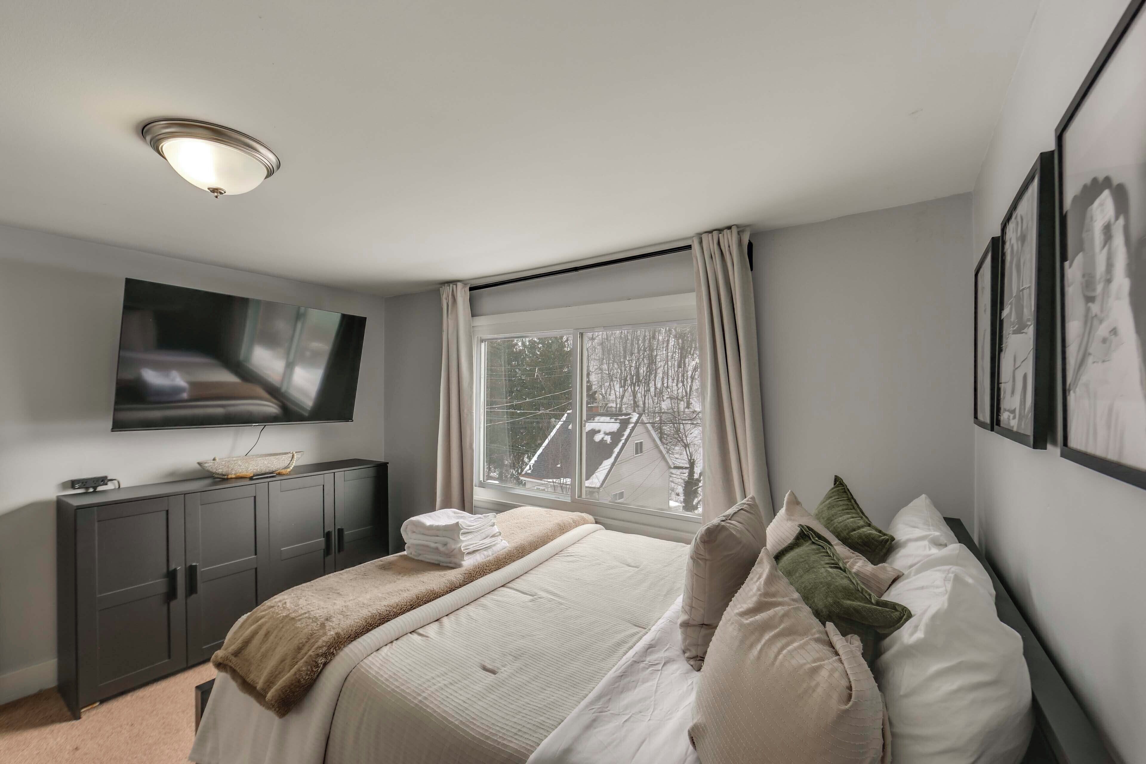 A bedroom with a bed, a TV mounted on the wall, a window with curtains, and a view of buildings outside.