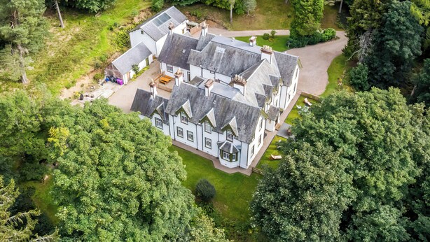 Eine Vogelperspektive auf ein großes, weißes, mehrstöckiges Haus mit gepflegtem Rasen und umgebendem Wald.