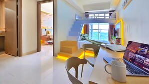A modern hotel room with a loft bed, a dining area with a laptop and mug, and a view of the outdoors. - Azora By SanLiving - 1BR - PIK (Jakarta)