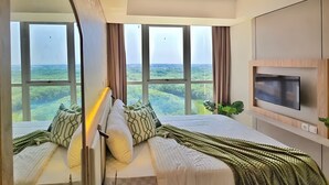 A bedroom with a large bed, a TV mounted on the wall, and a view of the landscape through large windows. - Azora By SanLiving - 1BR - PIK (Jakarta)