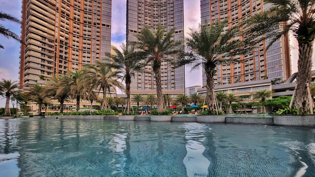 A swimming pool with clear water, surrounded by palm trees and tall buildings. - Azora By SanLiving - 1BR - PIK (Jakarta)