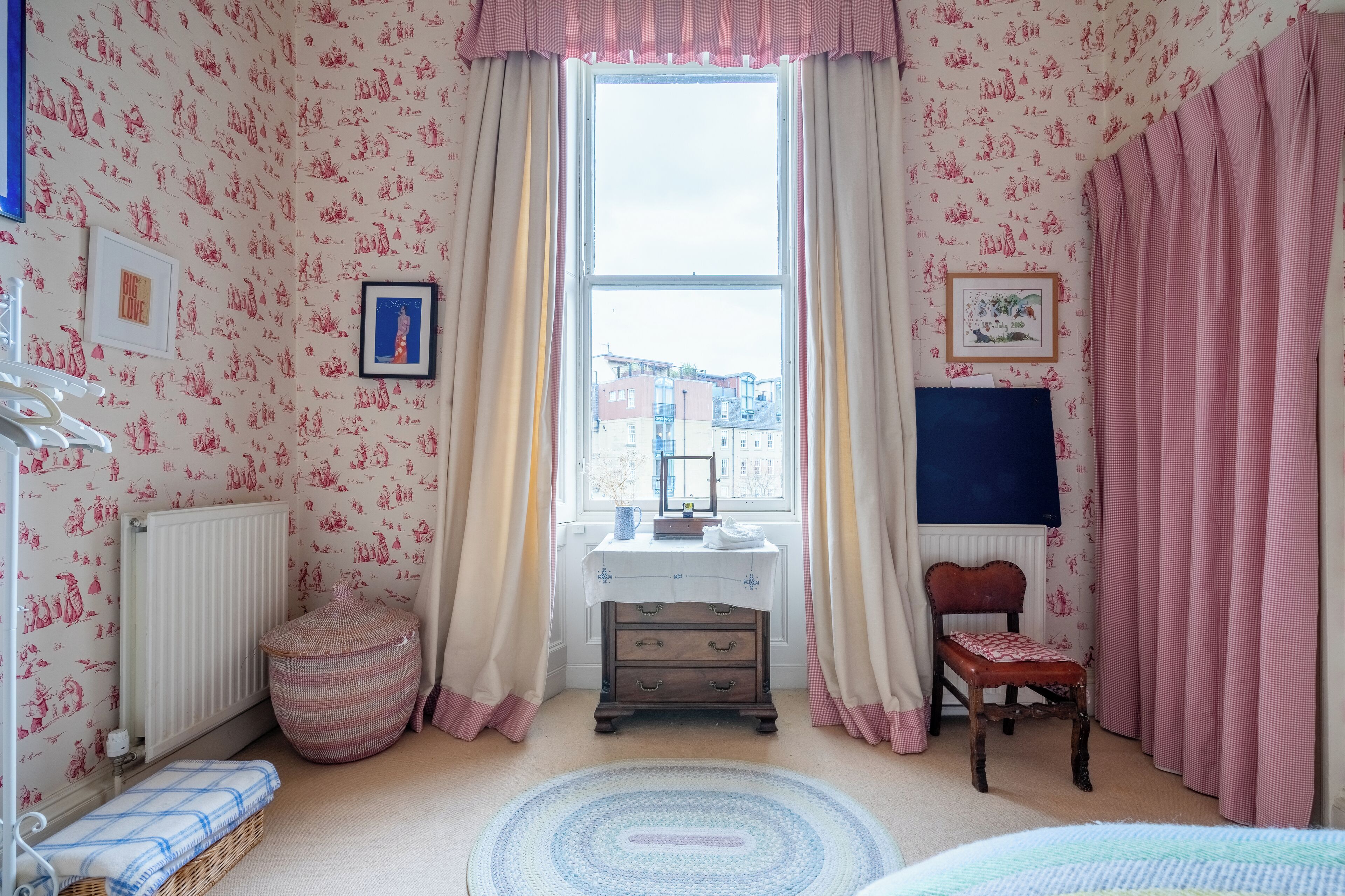 A child's bedroom with patterned wallpaper, a radiator, a desk with drawers, a chair, a window with curtains, and a bed with a blue and white checkered bedspread.