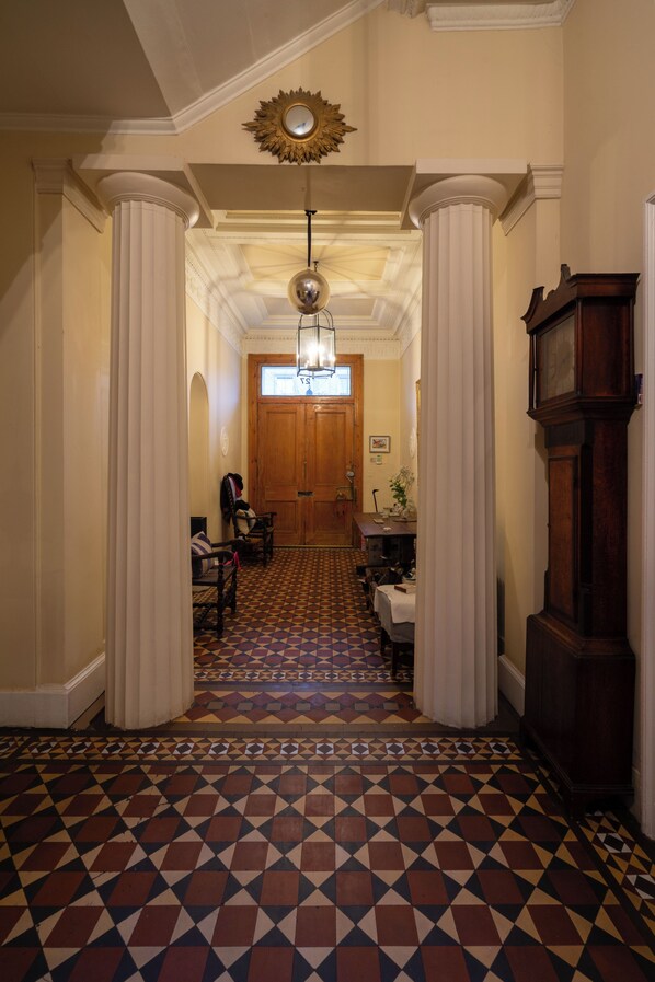A hallway with patterned flooring, white columns, a wooden door, and a grandfather clock.