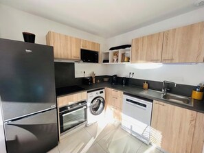 A modern kitchen with a black refrigerator, a washing machine, a dishwasher, wooden cabinets, and a microwave. - T3 large terrace - parking WIFI - CLIM 5P (La Grande-Motte)