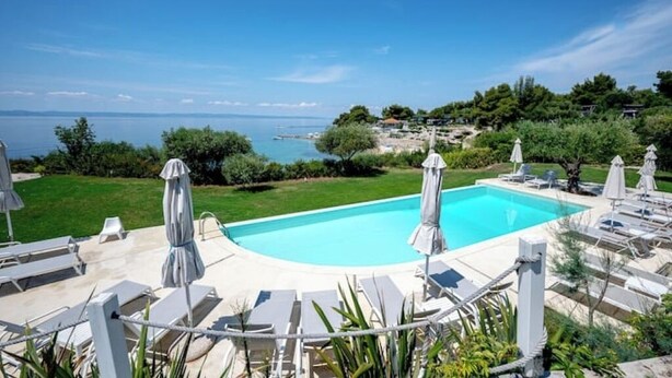 Piscina con sillas de descanso y sombrillas, rodeada de follaje y vista al mar.