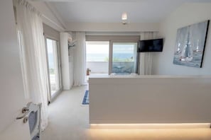 A modern hotel room with a large window offering a view of the sea, a flat-screen TV mounted on the wall, and a balcony with a table and chairs. - Villa Pathos: Beachfront Bliss with Pool & Panoramic Views/3 bdrs (Nikiti)