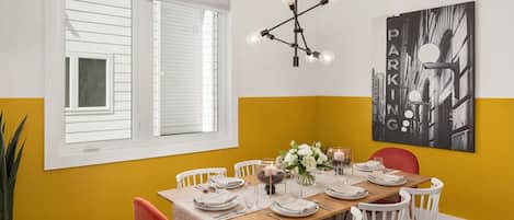Un comedor con una mesa de madera dispuesta para seis comensales, sillas blancas, una pared de acento amarilla, una obra de arte con un paisaje urbano en blanco y negro, y una lámpara moderna.