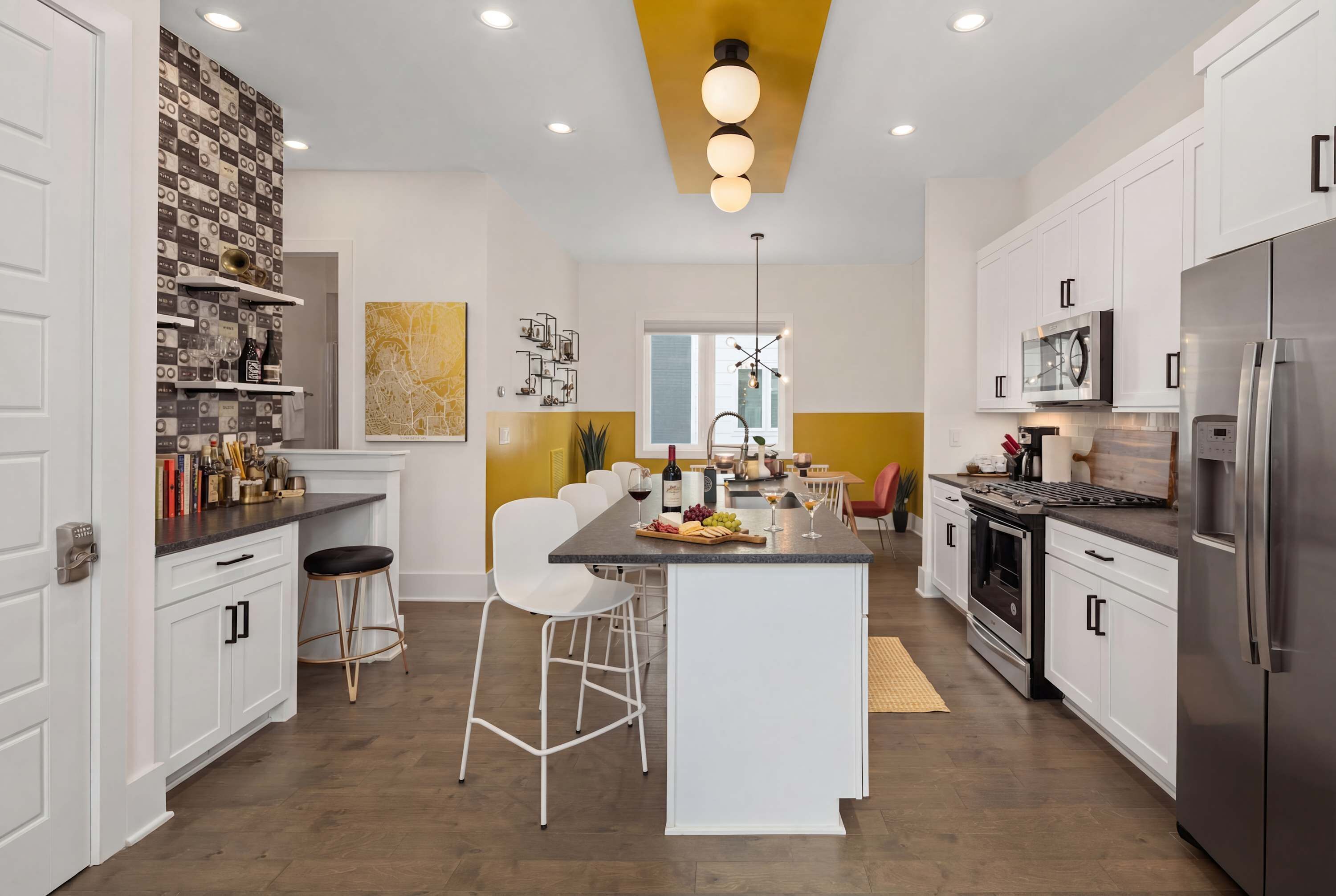Une cuisine moderne avec des meubles blancs, un îlot central, des tabourets de bar et un coin repas agrémenté d’un mur d’accent jaune.