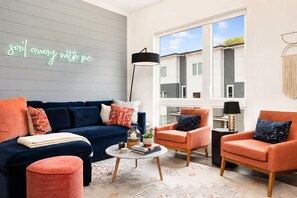 A modern living room with a blue sofa, orange armchairs, a coffee table, and a large window offering a view of buildings. - East Nashville weekend home with neon lounge, 3 king suites, sleeps 10 (Nashville)