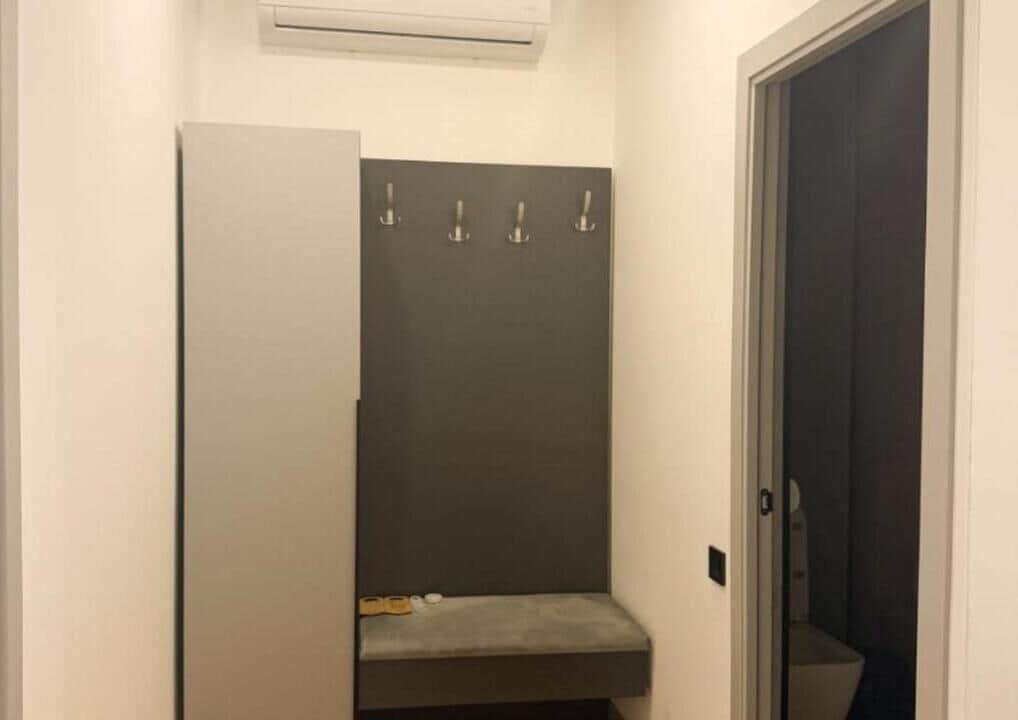 Ein schmaler Flur mit einer Tür rechts, einer Bank mit Handtuch und Schuh, sowie einer Wand-Klimaanlage.