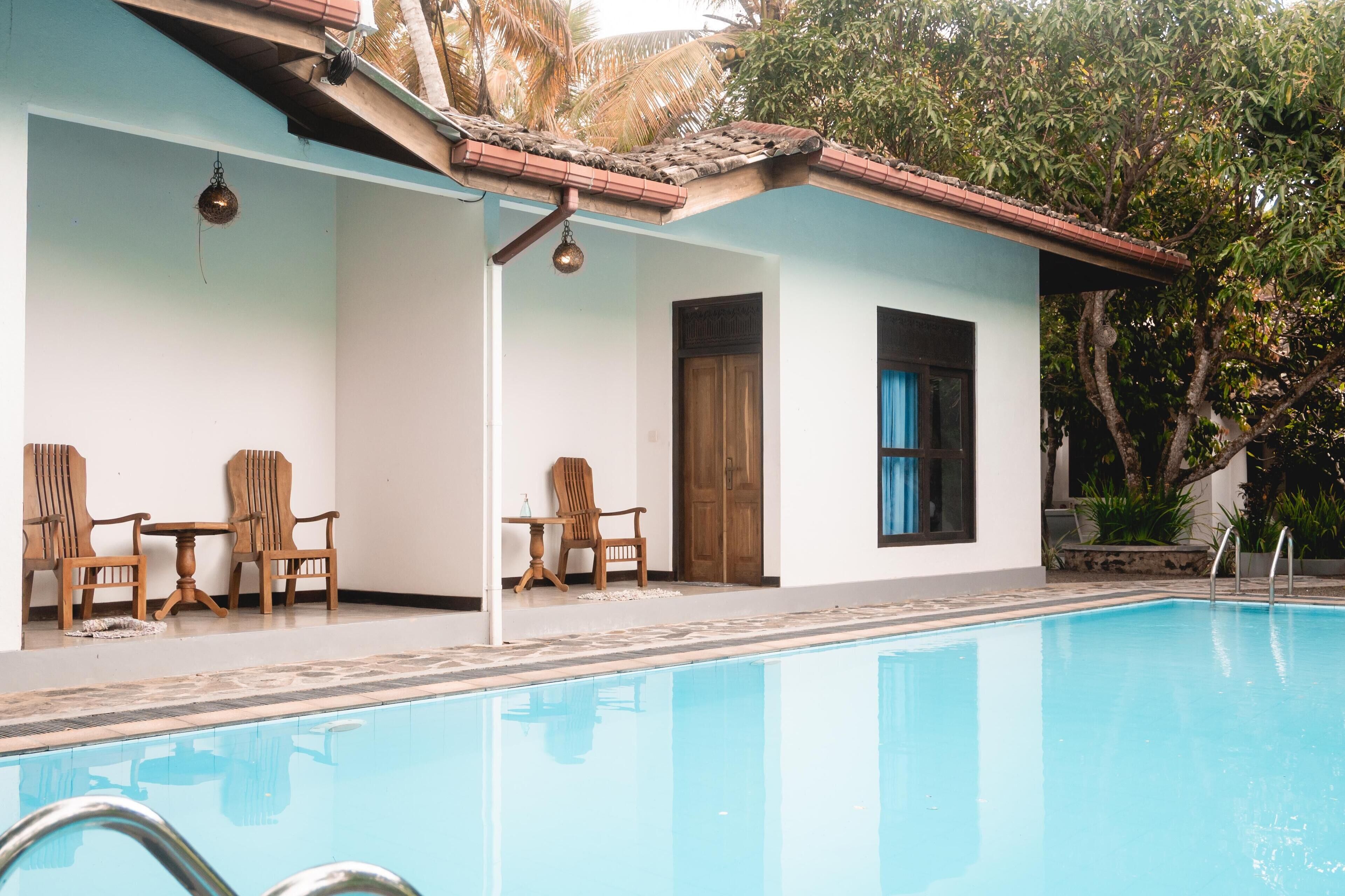 Una piscina con un bordeado de baldosas, sillas de madera y una zona de estar cubierta con una mesa y sillas.