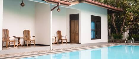 Una piscina con un bordeado de baldosas, sillas de madera y una zona de estar cubierta con una mesa y sillas.
