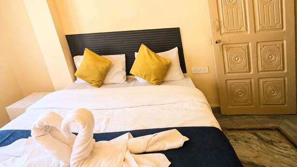 A neatly made bed with white linens and two yellow pillows, a dark headboard, and a wooden door with a geometric pattern. - GANGA KASHTI (Varanasi)