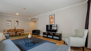 Un salon moderne comprenant un canapé, une table basse, un téléviseur posé sur un meuble noir, un coin repas avec des chaises et une table, et un fauteuil de couleur claire.
